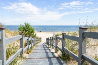 Eine flache Treppe mit hölzernem Geländer führt durch Sanddünen ans Meer. 