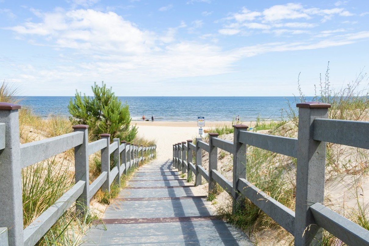 Eine flache Treppe mit hölzernem Geländer führt durch Sanddünen ans Meer. 