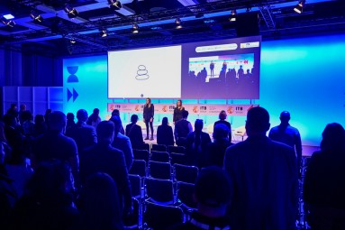 Two panellists stand on stage and speak to the audience.