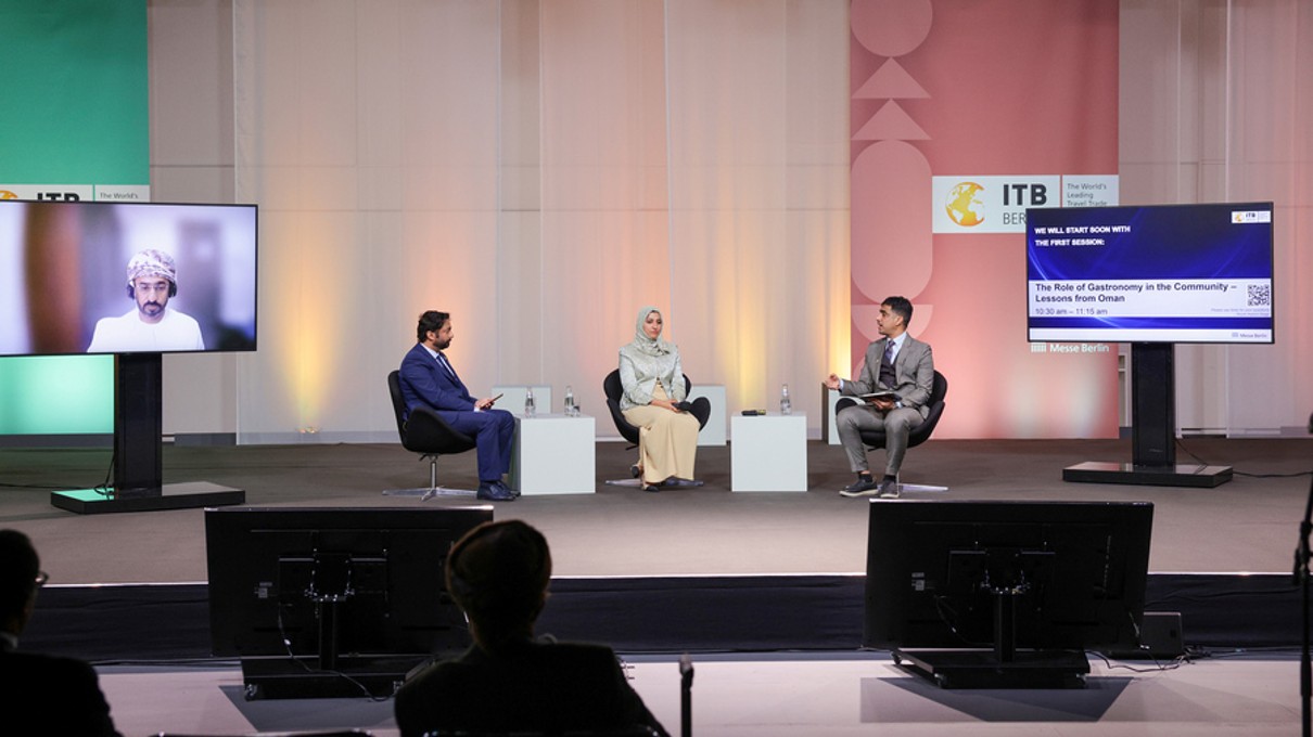 Three panellists speak on stage, another is digitally connected. 