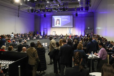 Das Bild zeigt eine große Konferenzhalle mit vielen Menschen, die in einem Halbkreis um eine zentrale Bühne sitzen