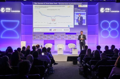 A man can be seen standing onstage, behind him a presentation, in the foreground the audience