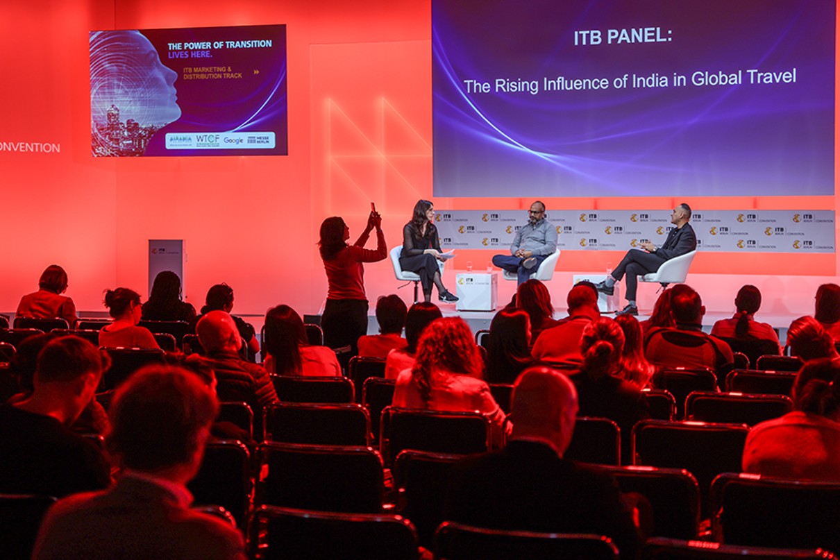 The image shows three people onstage having a discussion. The audience is in the foreground