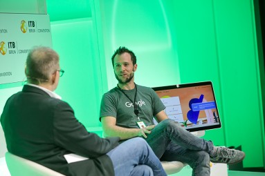 Two men are sitting on a stage and having a conversation, while a green-lit backdrop is visible in the background.