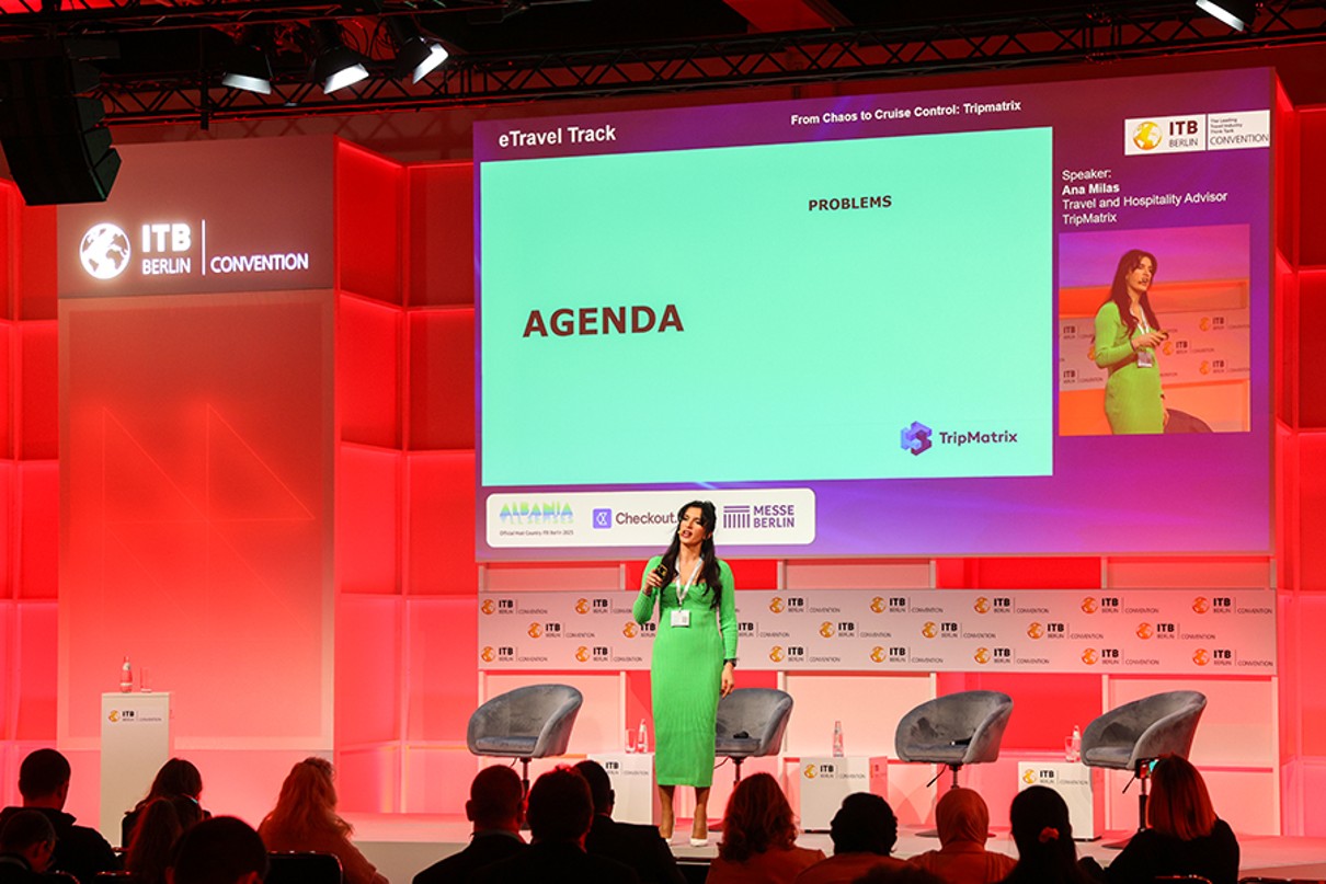 A woman in a green dress gives a presentation on a red-lit stage with a large screen behind her