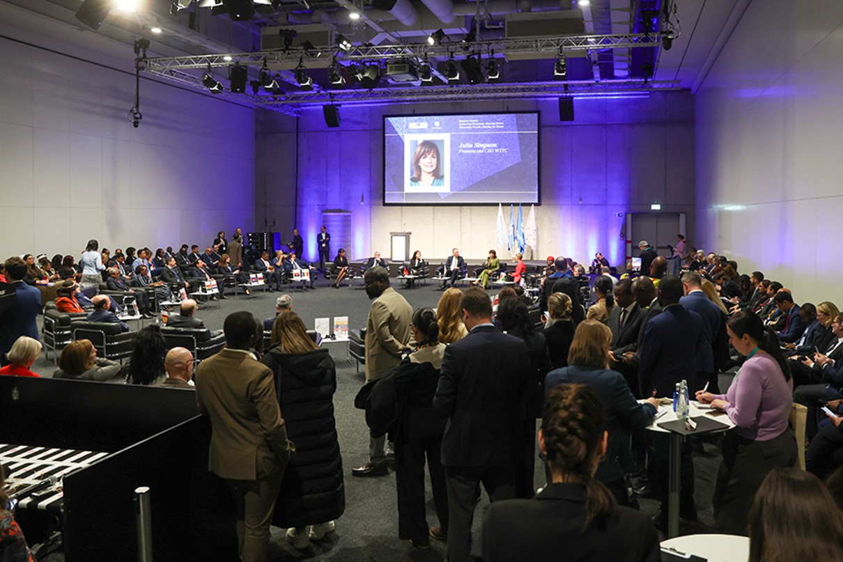 The image shows a large conference hall with many people seated in a semi-circle around a central stage