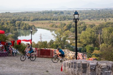 Two cyclists in a mountainous region in Albania, with a river and a green landscape in the background.