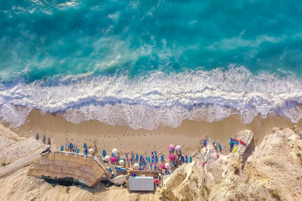 A bird's eye view of Pasqyra beach with its blue waters, sun umbrellas on the sandy beach and rocky mountains. 