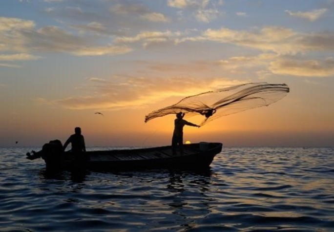 Ein Boot mit zwei Fischern auf dem Meer, ein Fischer wirft das Fischernetz aus. Am Horizont geht die Sonne unter.
