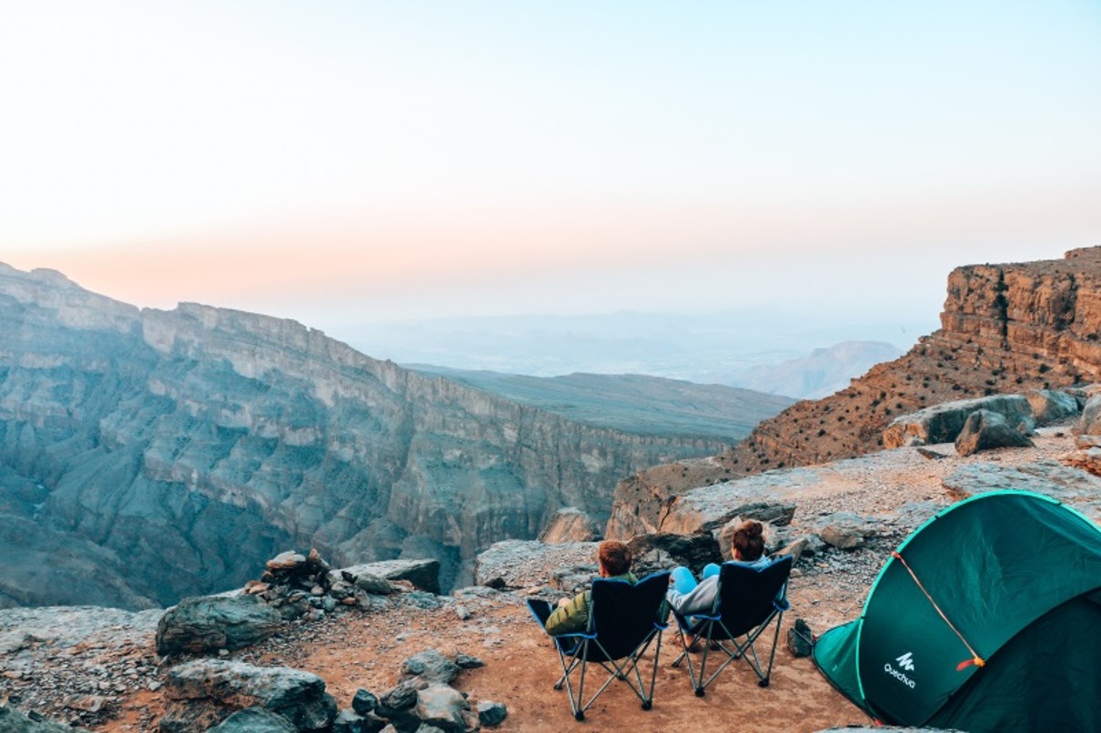 People sitting in front of a tent on a mountain, a valley in the background 
