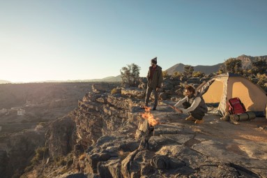 Two people on a mountain in front of a tent and campfire, the landscape of Jebel Akhdar in the Hajar Mountains in the background