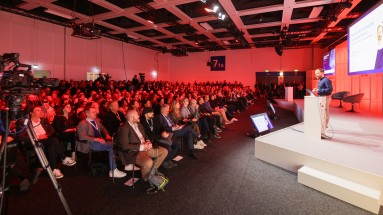 A packed hall with seated convention attendees listening to a lecture by a man on stage. 