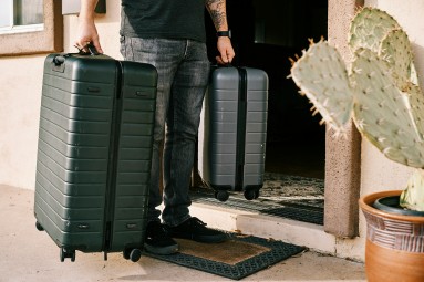 A man with a tattooed left arm is holding two black suitcases and standing next to an open front door. 