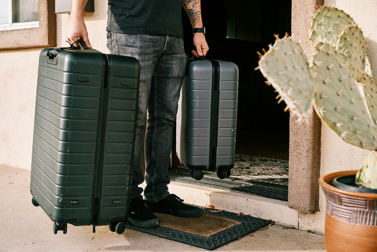 A man with a tattooed left arm is holding two black suitcases and standing next to an open front door. To the right in the foreground is a pot with a cactus plant. 