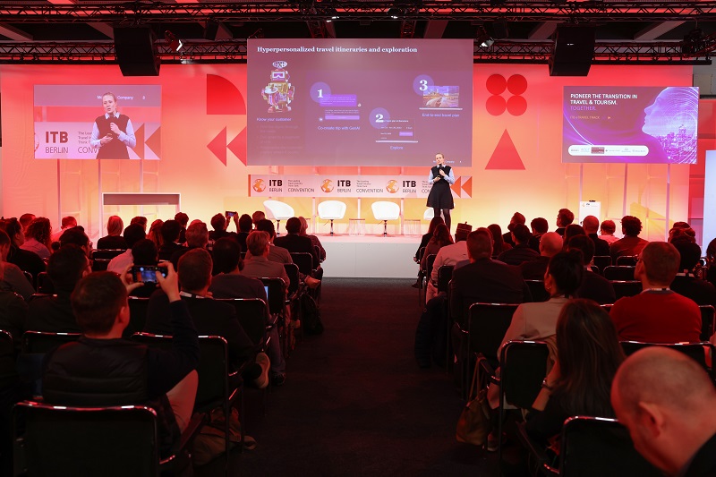 Audience in the foreground, directed towards a person on a stage holding a microphone. Audience in the foreground, directed towards a person on a stage holding a microphone.