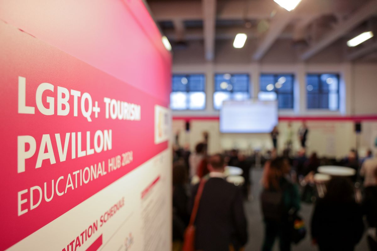 A red wall with LGBTQ+ Tourism Pavillon written on it in the foreground. In the background a stage with audience. A red wall with LGBTQ+ Tourism Pavillon written on it in the foreground. In the background a stage with audience.