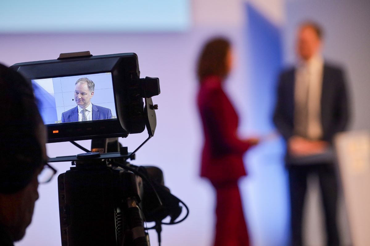 A camera is filming on stage. In the blury background you can see a women and a man talking. A camera is filming on stage. In the blury background you can see a women and a man talking.