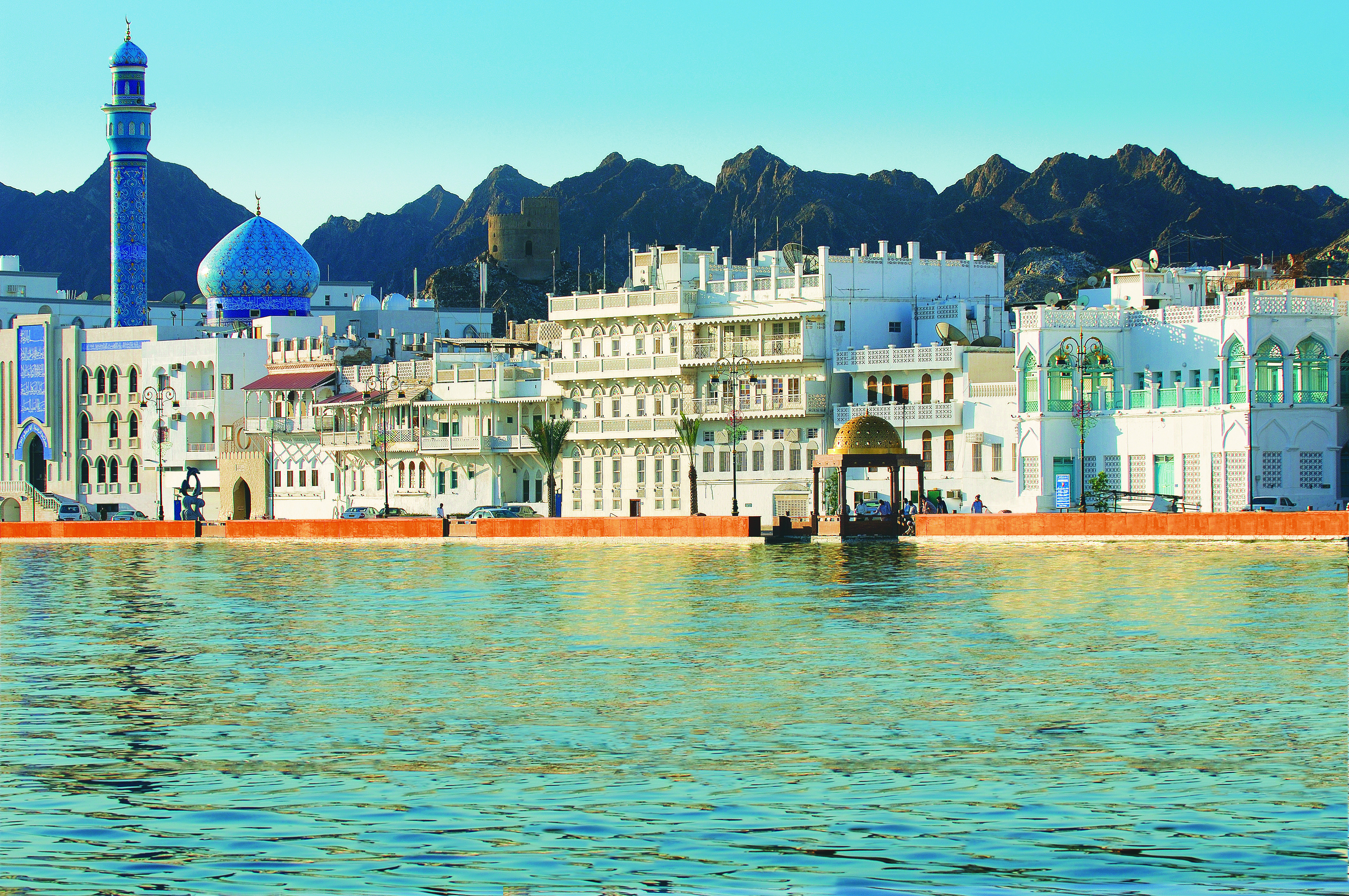 The Gulf of Oman is shown in the foreground. In the background is the waterfront Corniche of Muttrah with white houses and the Muttrah Mosque in Muscat, Oman. Behind it mountains are visible.